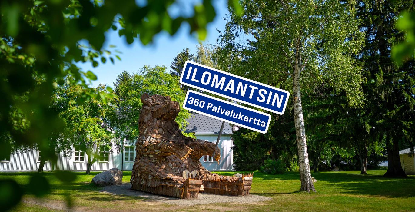 Suuri, puunpaloista koottu karhupatsas istuu tuolilla vehre&auml;ss&auml; puistossa valkoisen puutalon edess&auml;. Kuvan p&auml;&auml;ll&auml; on sininen kyltti, jossa lukee valkoisella "ILOMANTSIN 360 Palvelukartta".
