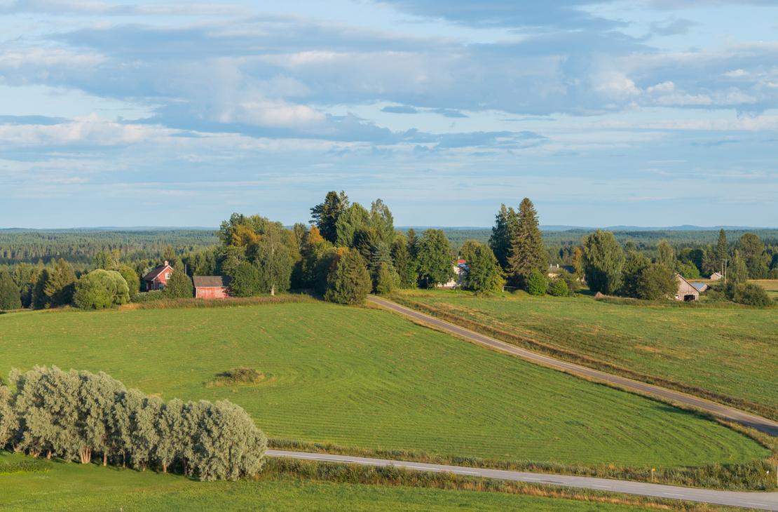 Ilmakuva kumpuilevasta peltomaisemasta ja sen läpi kulkevasta maantiestä. Rinteessä on punaisia ja valkoisia maalaistaloja sekä puurivistöjä.