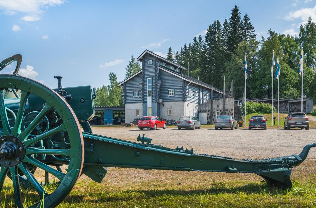 Kuva Taistelijan Talolta, joka esitt&auml;&auml; etualalla suurta, tummanvihre&auml;&auml;, vanhaa tykki&auml; tai haupitsia. Tykin suuri, puinen py&ouml;r&auml; n&auml;kyy etualalla vasemmalla. Taustalla on moderni, tummansininen tai harmaa museorakennus, jonka edess&auml; on hiekkaparkkipaikka ja joitakin autoja. Rakennuksen edess&auml; nousee useita lipputankoja, joissa liehuvat Suomen liput. Taivas on sininen ja pilvinen.