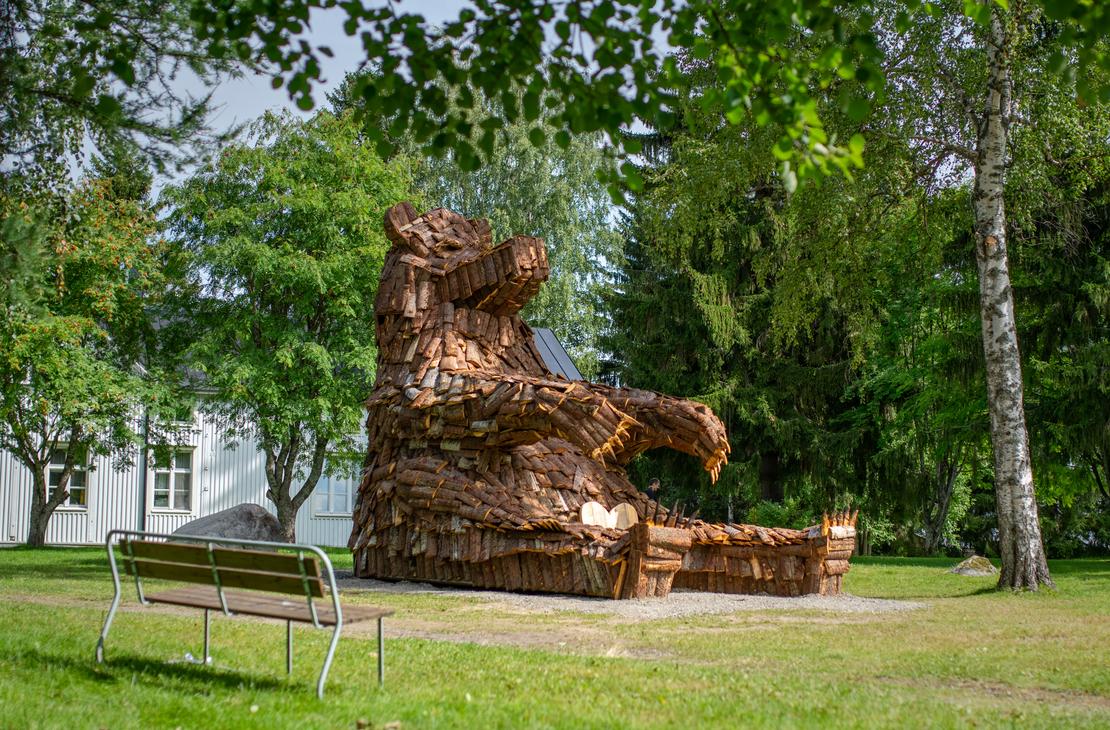 Lähikuva suuresta, puusta tehdystä karhupatsaasta, ILOKARHU -nimisestä veistoksesta Ilomantsista. Karhu on koottu useista pienemmistä puupaloista ja laudoista. Kuvassa näkyy selvästi karhun karkea pintakuvio. Taustalla on vihreää kasvillisuutta.