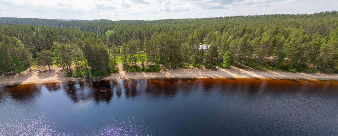 Panoraamailmakuva pitkästä hiekkarannasta, joka rajoittuu tiheään mäntymetsään. Aurinko heijastuu tumman järven pintaan ja taustalla siintää laaja metsämaisema.