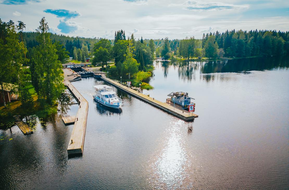 Ilmakuva tyynestä vesistöstä aurinkoisena päivänä, joka liittyy Blue Lake Cruisesin toiminta-alueeseen. Kuvassa näkyy kaksi venelaituria ja niihin kiinnitettynä useita aluksia, mukaan lukien suurempi, valkoinen risteilyalus ja pienempi vene. Laitureiden takana näkyy osa maata, mahdollisesti kanava tai sulku (vasemmalla), ja aluetta ympäröi tiheä, vehreä metsä. Auringonvalo heijastuu kirkkaasti veden pintaan oikealla.