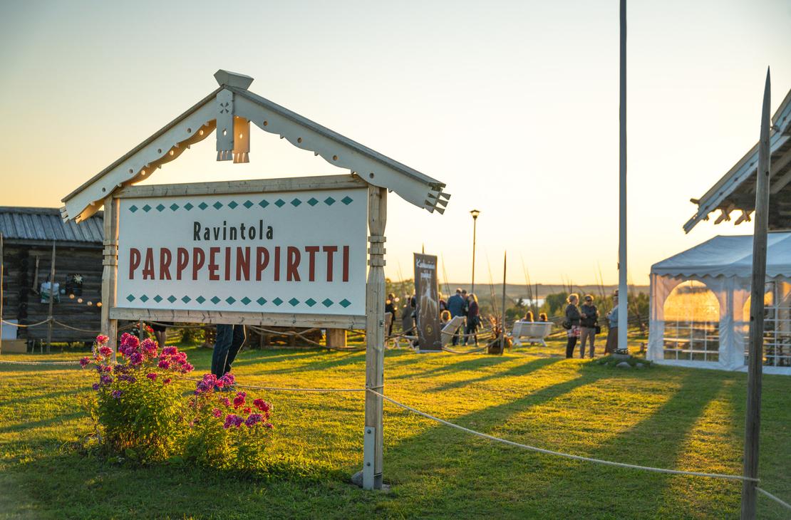 Lähikuva puisesta "Ravintola Parppeinpirtti" -kyltistä ilta-auringon paisteessa. Etualalla on kukkia, ja taustalla näkyy ihmisiä ravintolan pihapiirissä.