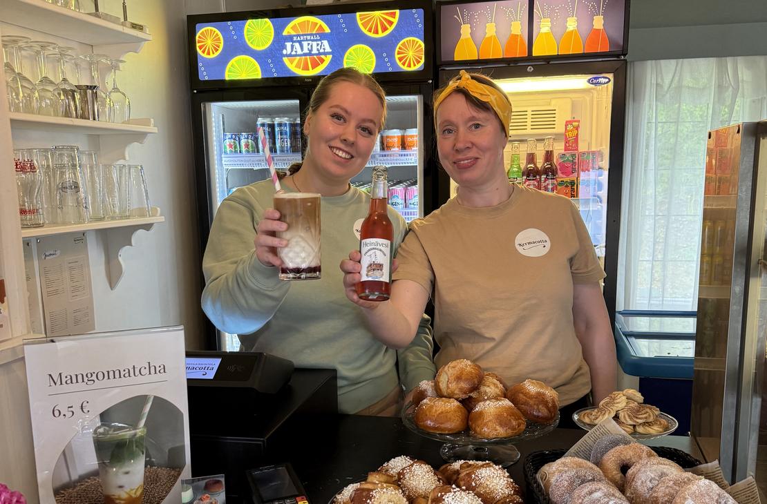 Kaksi hymyilevää työntekijää Satamakahvila-ravintola Kermacotan tiskillä. Toisella työntekijällä on käsissään pullollinen juomaa ja toisella täytetty lasi. Tiskin päällä on esillä houkuttelevia, vastaleivottuja korvapuusteja ja muita leivonnaisia koreissa. Taustalla näkyy kylmäkaappeja juomille.