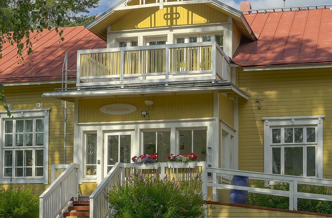 Lähikuva Äksyt Ämmät -yrityksen rakennuksesta, joka on keltainen, perinteinen puutalo punaisella harjakatolla. Rakennuksessa on toisessa kerroksessa pieni, valkokaiteinen parveke ja alakerrassa valkoiset ikkunankarmit. Kuvan edessä näkyy valkoista aitaa tai ramppia. Rakennusta reunustaa vasemmalla vihreät puut.