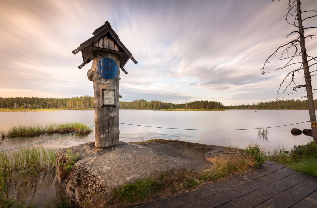 Kuva Suomen itäisimmältä pisteeltä auringonlaskun tai -nousun aikaan. Etualalla on puinen, veistetty muistomerkki tai pylväs, jonka päällä on pieni katos ja sininen, pyöreä symboli. Muistomerkki seisoo kivetyllä alustalla veden äärellä. Taustalla avautuu laaja järvimaisema, jonka vastarannalla on tummaa metsää. Taivas on dramaattinen, pilvinen, ja siinä näkyy pitkän valotusajan aiheuttamaa liikettä. Oikealla näkyy kuollut, karu puu ja osittain puinen kävelysilta.