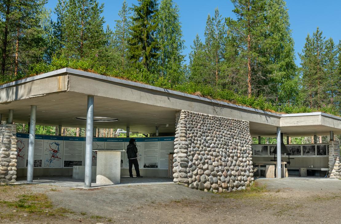 Kuva &Ouml;ykk&ouml;senvaaran taistelualueen taukopaikasta tai museorakennuksesta. Se on moderni, matala, osittain avoin katos, jossa on vihre&auml;, maanpeitt&auml;m&auml; katto. Rakenne koostuu betonista ja py&ouml;reill&auml; luonnonkivill&auml; verhoilluista pylv&auml;ist&auml;. Rakennuksen alla on taukopaikka tai n&auml;yttelytila. Rakennus sijaitsee mets&auml;n reunassa soraisella maapohjalla.