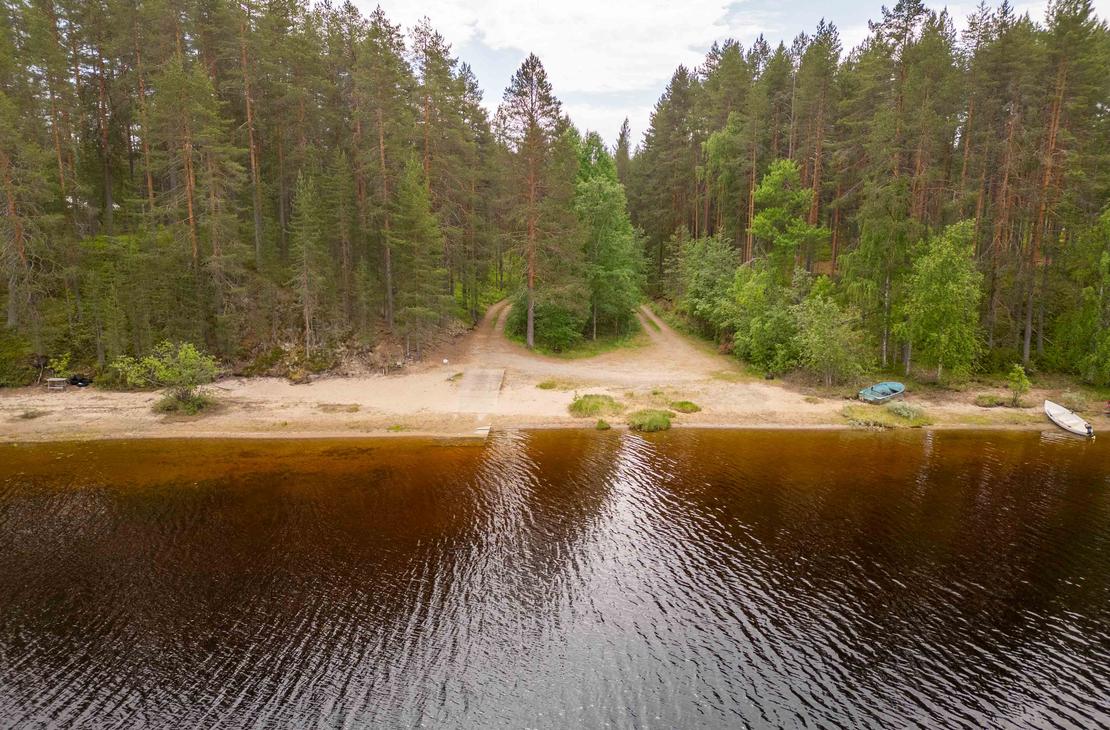 Ilmakuva järven hiekkarannasta ja veneenlaskupaikasta. Rantaan johtaa kaksi hiekkatietä tiheän mäntymetsän läpi, ja rantavedessä kimmeltää tumma vesi.