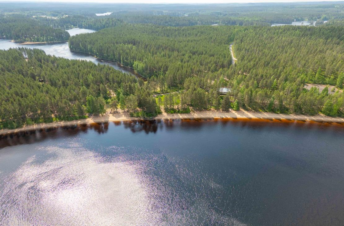 Korkealta otettu laaja panoraamailmakuva Suomen järvi- ja metsämaisemasta. Etualalla on kaareva hiekkaranta ja sen takana silmänkantamattomiin jatkuvaa tiheää havumetsää ja vesistöjä.