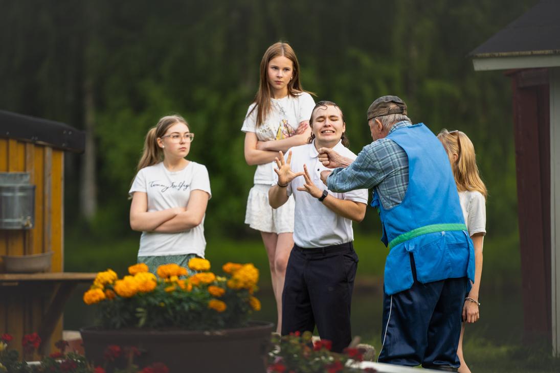 Möhkön kesäteatterin Peitetarina-näytelmän kohtaus, jossa siniseen liiviin pukeutunut vanhempi mies elehtii lähellä valkoiseen poolopaitaan pukeutunutta nuorta miestä, joka tekee torjuvan käsieleen kasvot hätääntyneenä. Taustalla kolme nuorta naista seuraa tilannetta vakavina, ja etualalla ruukussa kukkii oransseja samettikukkia.