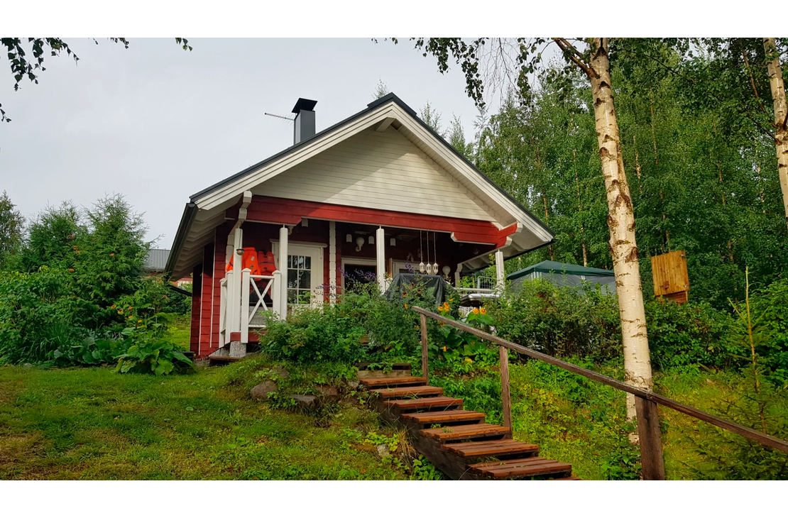 Kuva esittää Tujanranta B&B:n pientä, perinteistä punamultamaalilla maalattua mökkiä tai aittarakennusta. Rakennuksessa on valkoiset ikkunankarmit ja päätykolmio sekä tumma harjakatto. Mökin edessä on rinteessä puiset portaat ja kaiteet. Rakennusta ympäröi runsas, vihreä kasvillisuus ja koivuja.