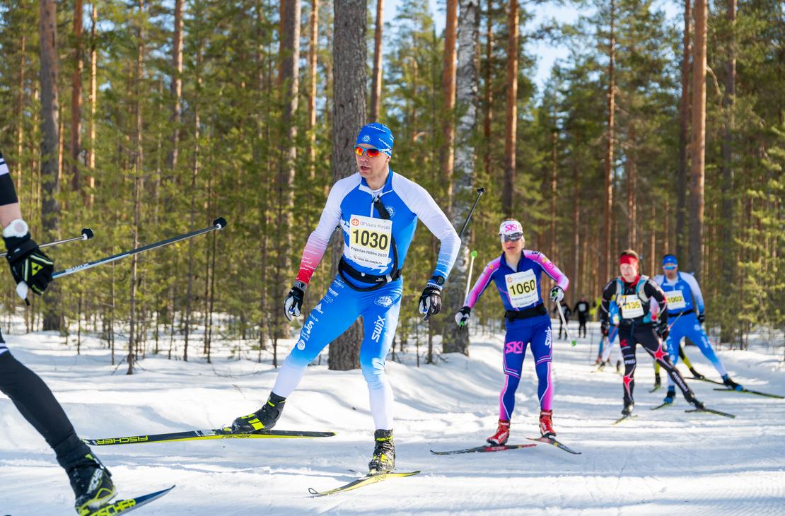 uva hiihtokilpailusta aurinkoisessa, lumisessa mets&auml;maastossa. Etualalla useita hiiht&auml;ji&auml; on liikkeess&auml;. Keskeisen&auml; hahmona on mies, jolla on sinivalkoinen, numerolla 1030 varustettu hiihtoasu, ja h&auml;n k&auml;ytt&auml;&auml; luistelutyyli&auml;. H&auml;nen takanaan on nainen pinkin- ja sinis&auml;vyisess&auml; asussa, numerolla 1060, ja muita hiiht&auml;ji&auml; kauempana. Maasto on tasaista, ja taustalla on tihe&auml;, m&auml;ntyvaltainen mets&auml;.