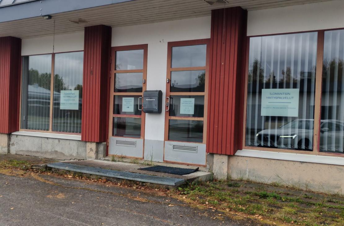 Ilomantsin Yrityspalvelut -toimiston sisäänkäynti ulkoapäin. Yksikerroksisen rakennuksen julkisivussa on valkoista seinää, pystysuuntaisia punaruskeita puupaneeleita sekä suuria ikkunoita pystylamellikaihtimilla. Keskellä on kaksi ruskeakehyksistä lasiovea, joiden välissä on harmaa postilaatikko ja edessä metalliritiläinen askelma mattoineen. Oikeanpuoleisessa ikkunassa on valkopohjainen kyltti, jossa lukee "ILOMANTSIN YRITYSPALVELUT KIRJANPITO - PALKANLASKENTA - VEROASIAT". Etualalla on asfaltoitu piha-alue, ja rakennuksen seinustalla kasvaa hieman ruohoa.