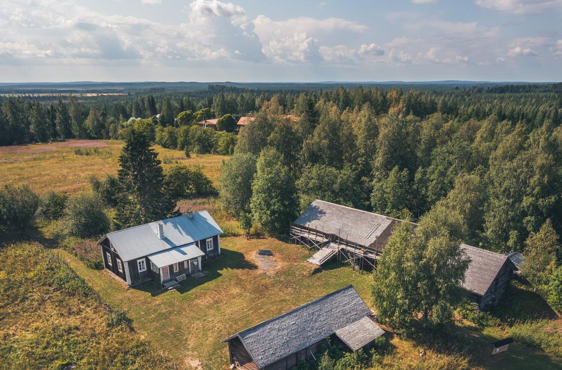 Ilmakuva Makkolan museotilasta, joka koostuu useista perinteisistä, tummista hirsirakennuksista harmailla katoilla. Rakennukset sijaitsevat vihreällä niityllä, jota ympäröi tiheä metsä ja aukea pelto. Piha-alueella näkyy ajopolkuja ja pieni avoin alue rakennusten välissä.