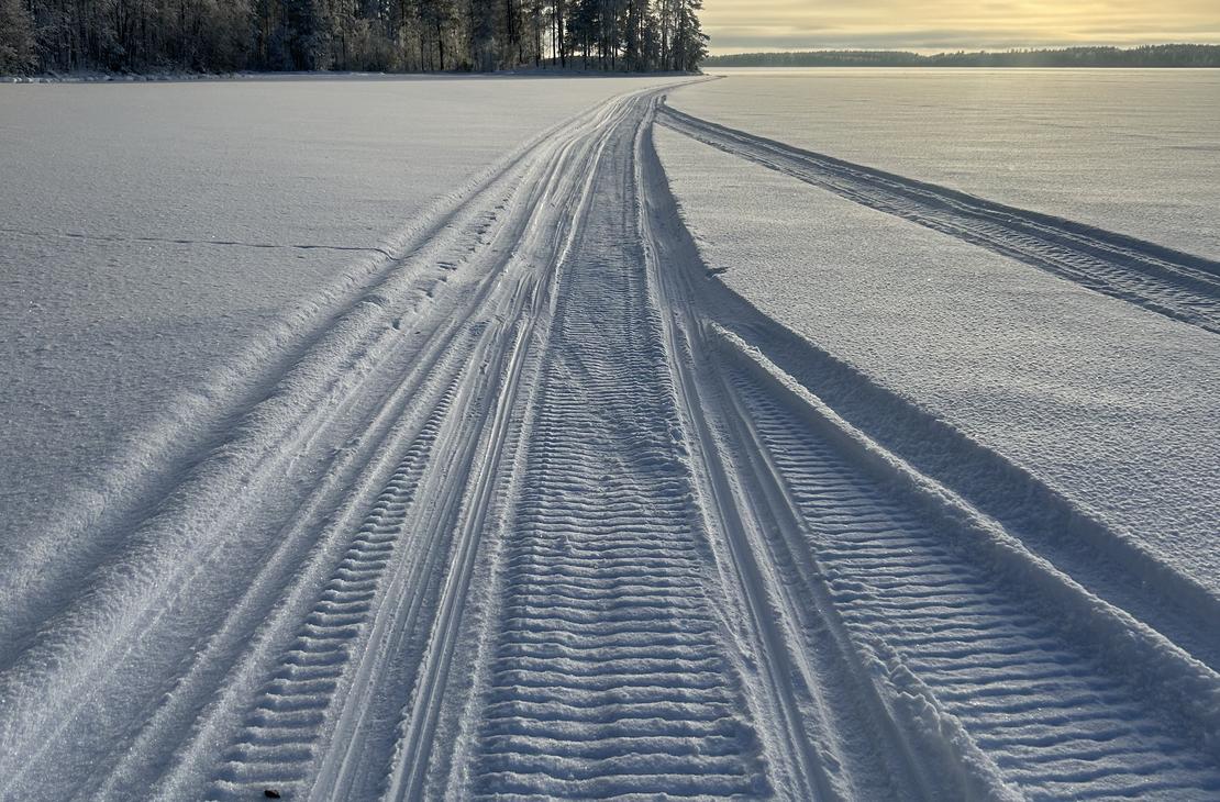 Perspektiivikuva Moottorikelkka urasta, joka kulkee lumisella järven jäällä kohti matalalla paistavaa aurinkoa ja kaukana häämöttävää metsänreunaa.