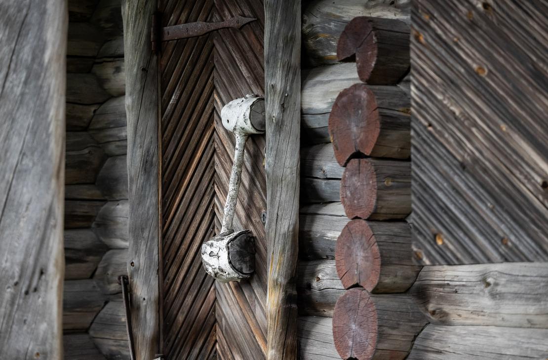 Yksityiskohta vanhan harmaan hirsirakennuksen ovesta, jossa on vinoon asetetut puulaudat ja luonnonmuotoisesta koivunoksasta valmistettu vedin. Ruosteinen saranarauta ja kirveell&auml; veistetyt hirrenp&auml;&auml;t n&auml;kyv&auml;t oven vieress&auml;.