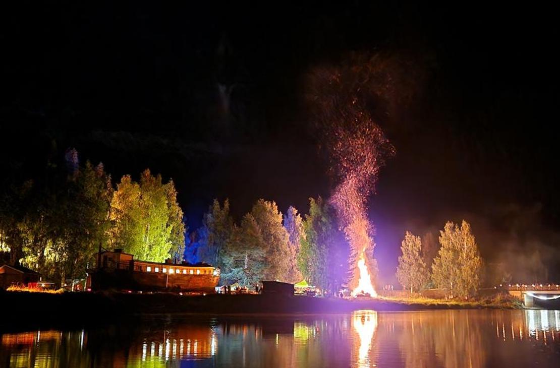 Yökuva Möhköstä vesistön äärellä järjestetystä tapahtumasta. Keskellä hehkuu suuri, korkea kokko tai roihuava nuotio, jonka liekit valaisevat taivaan. Kokko heijastuu kirkkaana tyyneen veteen etualalla. Rantaviivan puita on valaistu erivärisillä jouluvaloilla, luoden keltaisia, vihreitä ja violetteja sävyjä pimeään. Taustalla näkyy rakennuksen siluetti.