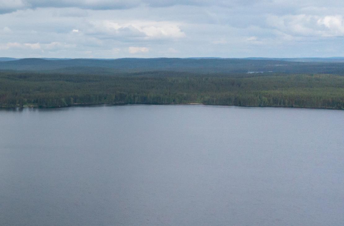 Laaja panoraamakuva, joka on otettu korkealta. Se esittää tyyntä, tummansinistä järven pintaa, joka täyttää suurimman osan kuvasta. Järven takana horisontissa näkyy tiheä, tummanvihreä metsä ja taustalla kaukaisia metsäisiä kukkuloita. Taivas on pilvinen ja vaaleanharmaa.