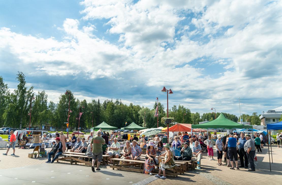 Ihmisiä istumassa puupenkeillä ja kiertelemässä Ilomantsin torilla värikkäiden myyntitelttojen välissä aurinkoisena päivänä suuren, valkopilvisen taivaan alla.