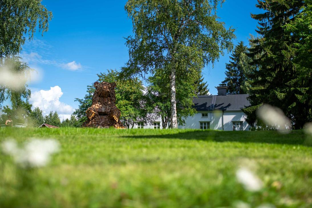 Laajakuva suuresta, puusta tehdystä karhupatsaasta, ILOKARHU -nimisestä veistoksesta Ilomantsista. Karhu on koottu useista pienemmistä puupaloista ja laudoista. Kuvassa näkyy selvästi karhun karkea pintakuvio. Taustalla on vihreää kasvillisuutta ja sininen taivas. Etualalla on muutama valkoinen kukka.
