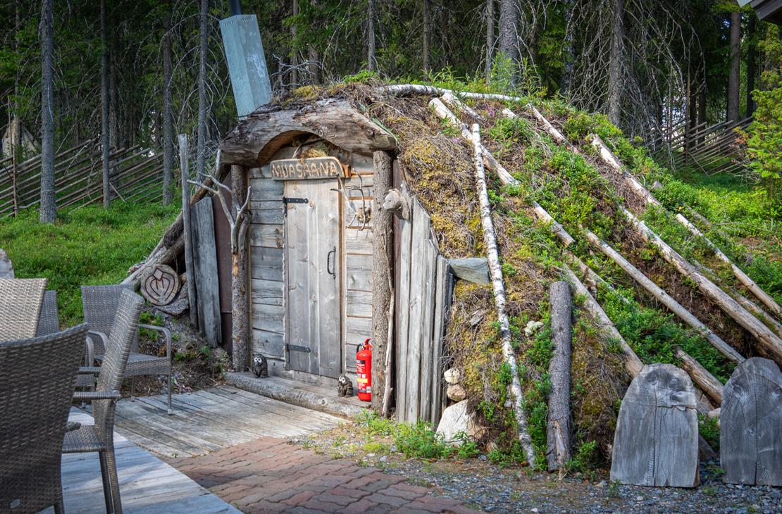 Maahan osittain upotettu, turvekattoinen "MUASAANA". Rakennuksen sein&auml;t on tuettu kelopuin ja oven vieress&auml; on punainen vaahtosammutin sek&auml; pieni&auml; p&ouml;ll&ouml;veistoksia.