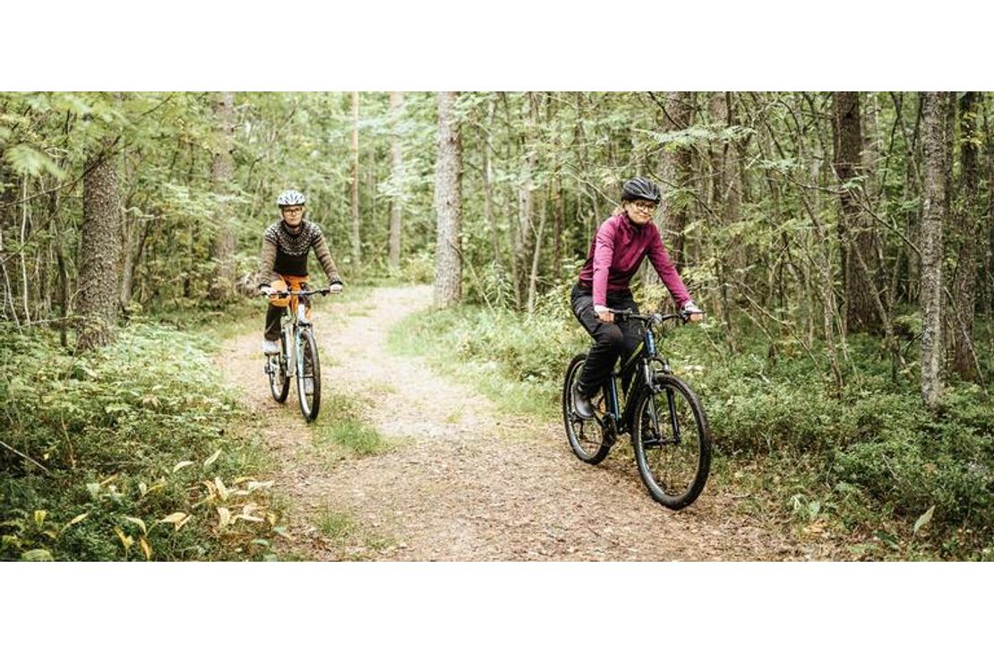 Kaksi henkilöä, nainen etualalla ja toinen henkilö taustalla, pyöräilevät mutkittelevaa hiekkatietä pitkin tiheässä metsässä. Nainen etualalla on pukeutunut pinkkiin paitaan ja kypärään, ja hän ajaa tummaa maastopyörää. Taustalla oleva pyöräilijä on pukeutunut tummiin vaatteisiin ja ajaa vaaleampaa pyörää. Polkua reunustaa runsas, vihreä aluskasvillisuus.