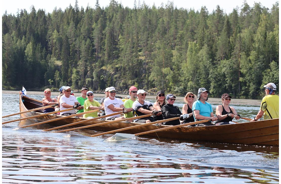 Pitkä puinen kirkkovene täynnä soutajia lipuu tyynellä järvellä. Taustalla näkyy jyrkkä metsäinen rinnerynnäs.