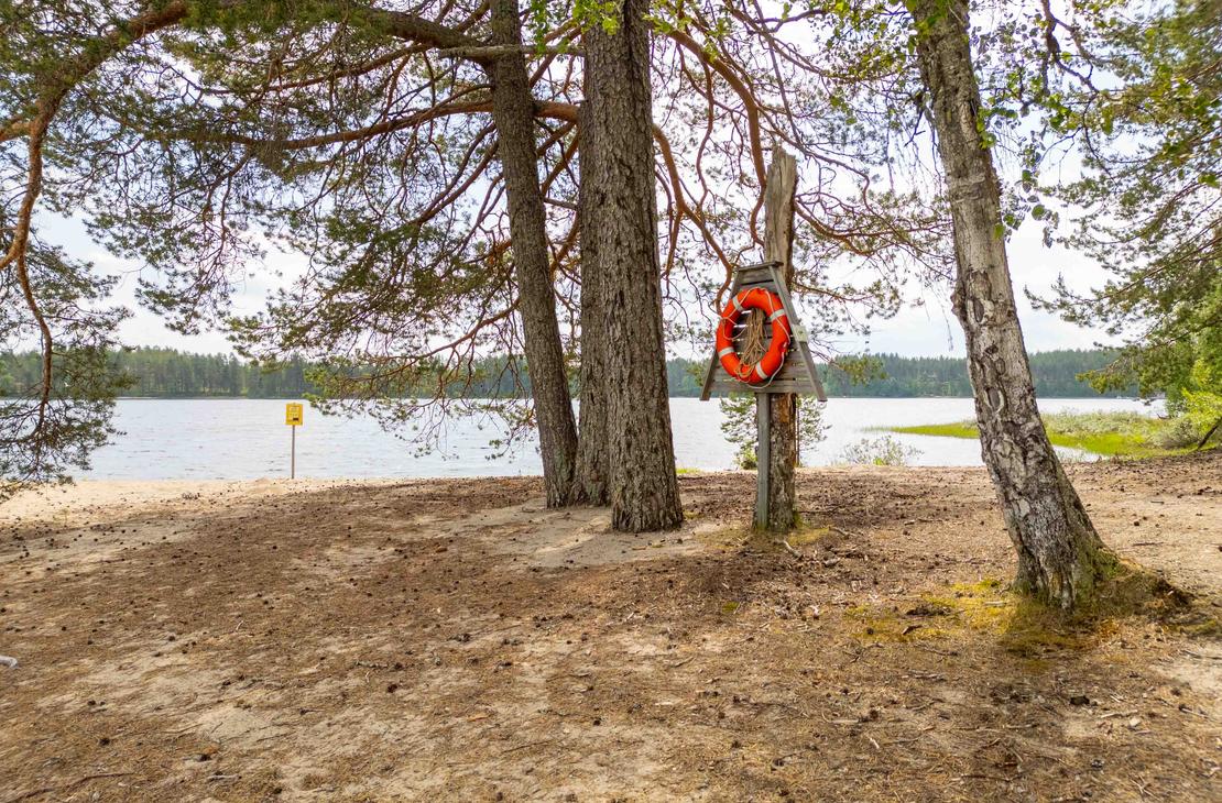 Punavalkoinen pelastusrengas kiinnitettynä puiseen telineeseen rantamännikön siimeksessä. Taustalla avautuu vaalea hiekkaranta ja tyyni järvimaisema.