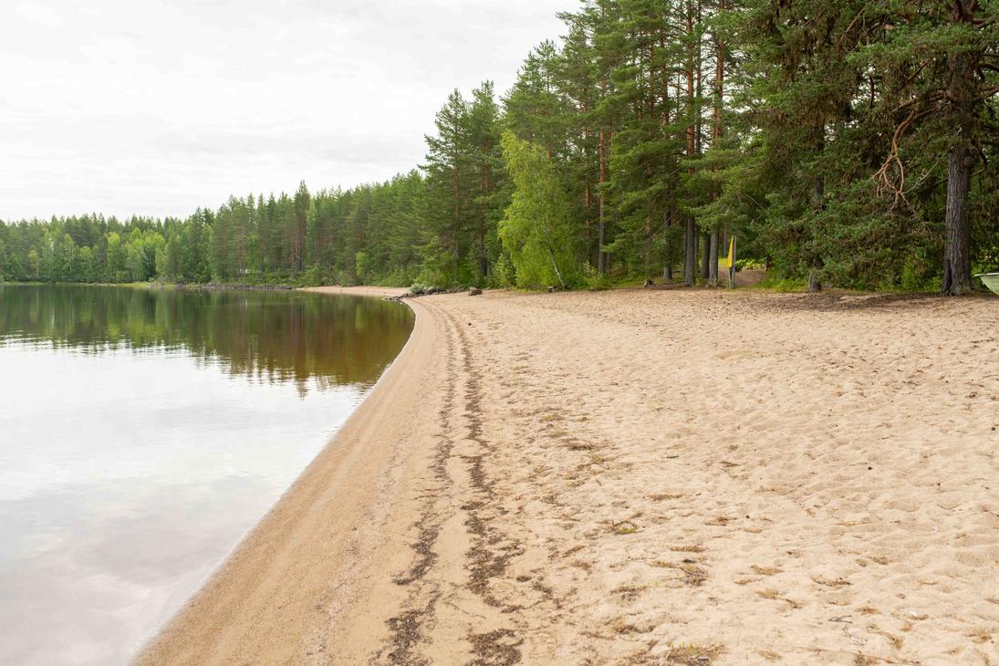 Pitkä hiekkaranta järven rannalla. Rannan vieressä kasvaa tiheä mäntymetsä, ja tyyni vedenpinta peilaa vastarannan puita.