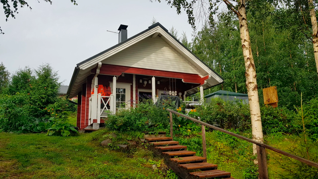 Kuva esittää Tujanranta B&B:n pientä, perinteistä punamultamaalilla maalattua mökkiä tai aittarakennusta. Rakennuksessa on valkoiset ikkunankarmit ja päätykolmio sekä tumma harjakatto. Mökin edessä on rinteessä puiset portaat ja kaiteet. Rakennusta ympäröi runsas, vihreä kasvillisuus ja koivuja.