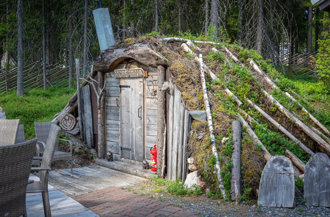Maahan osittain upotettu, turvekattoinen "MUASAANA". Rakennuksen sein&auml;t on tuettu kelopuin ja oven vieress&auml; on punainen vaahtosammutin sek&auml; pieni&auml; p&ouml;ll&ouml;veistoksia.