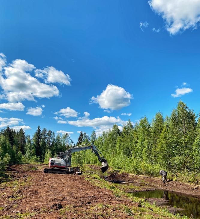 Takeuchi kaivinkone kaivaa kosteikkoallasta. Aurinkoinen kes&auml;p&auml;iv&auml; ja puut vihert&auml;v&auml;t kaivinkoneen ymp&auml;rill&auml;.