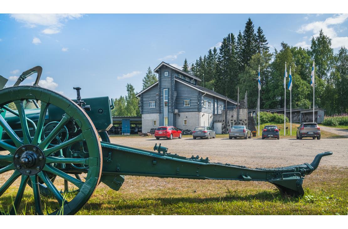 Kuva Taistelijan Talolta, joka esitt&auml;&auml; etualalla suurta, tummanvihre&auml;&auml;, vanhaa tykki&auml; tai haupitsia. Tykin suuri, puinen py&ouml;r&auml; n&auml;kyy etualalla vasemmalla. Taustalla on moderni, tummansininen tai harmaa museorakennus, jonka edess&auml; on hiekkaparkkipaikka ja joitakin autoja. Rakennuksen edess&auml; nousee useita lipputankoja, joissa liehuvat Suomen liput. Taivas on sininen ja pilvinen.