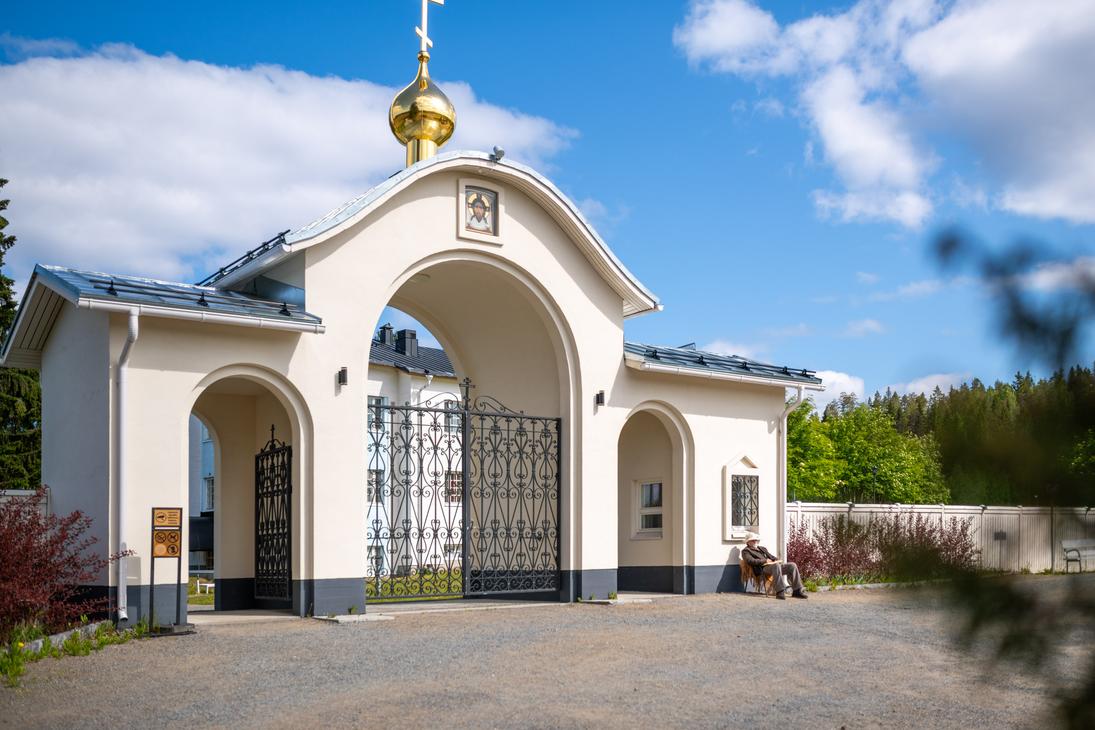 Valamon luostarin vaalea, kaari-aukkoinen porttirakennus, jonka yläpuolella on kultainen sipulikupoli ja risti. Koristeellinen, musta metalliportti on suljettu. Oikealla rakennuksen seinustalla istuu henkilö penkillä aurinkoisena päivänä.