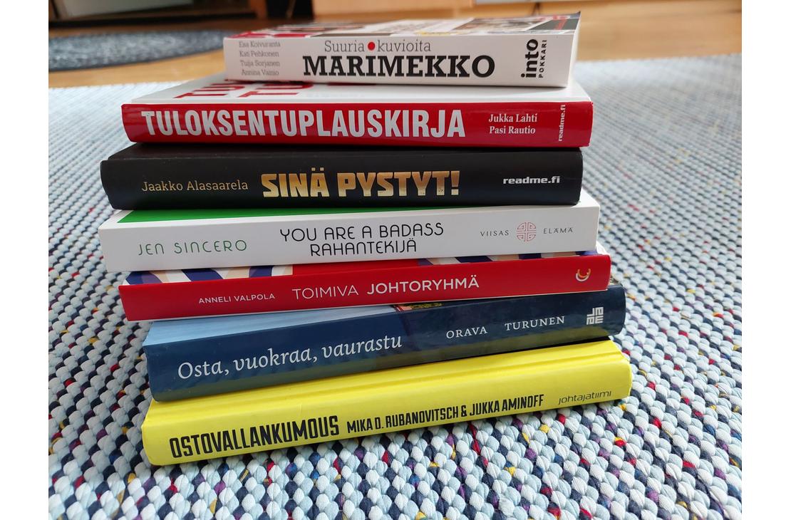 kuvassa on pino kirjoja, jotka on pinottu päällekkäin. Kirjojen kannet ovat erivärisiä ja -kuvioisia, ja niissä on erilaisia tekstejä. Pinon päällimmäinen kirja on vaaleansininen ja siinä lukee "MARIMEKKO". Pinon alimmainen kirja on keltainen ja siinä lukee "OSTA VAIN, JOS TARVITSET". Muissa kirjoissa on tekstejä kuten "TULOKSENTUPLAUSKIRJA", "SINÄ PYSTYT!", "YOU ARE A BADASS", "JOHTAJA ILMAN JOUKKOJA", "OMA RAHA, OMAT SÄÄNNÖT". Kirjat on asetettu harmaalle matolle.