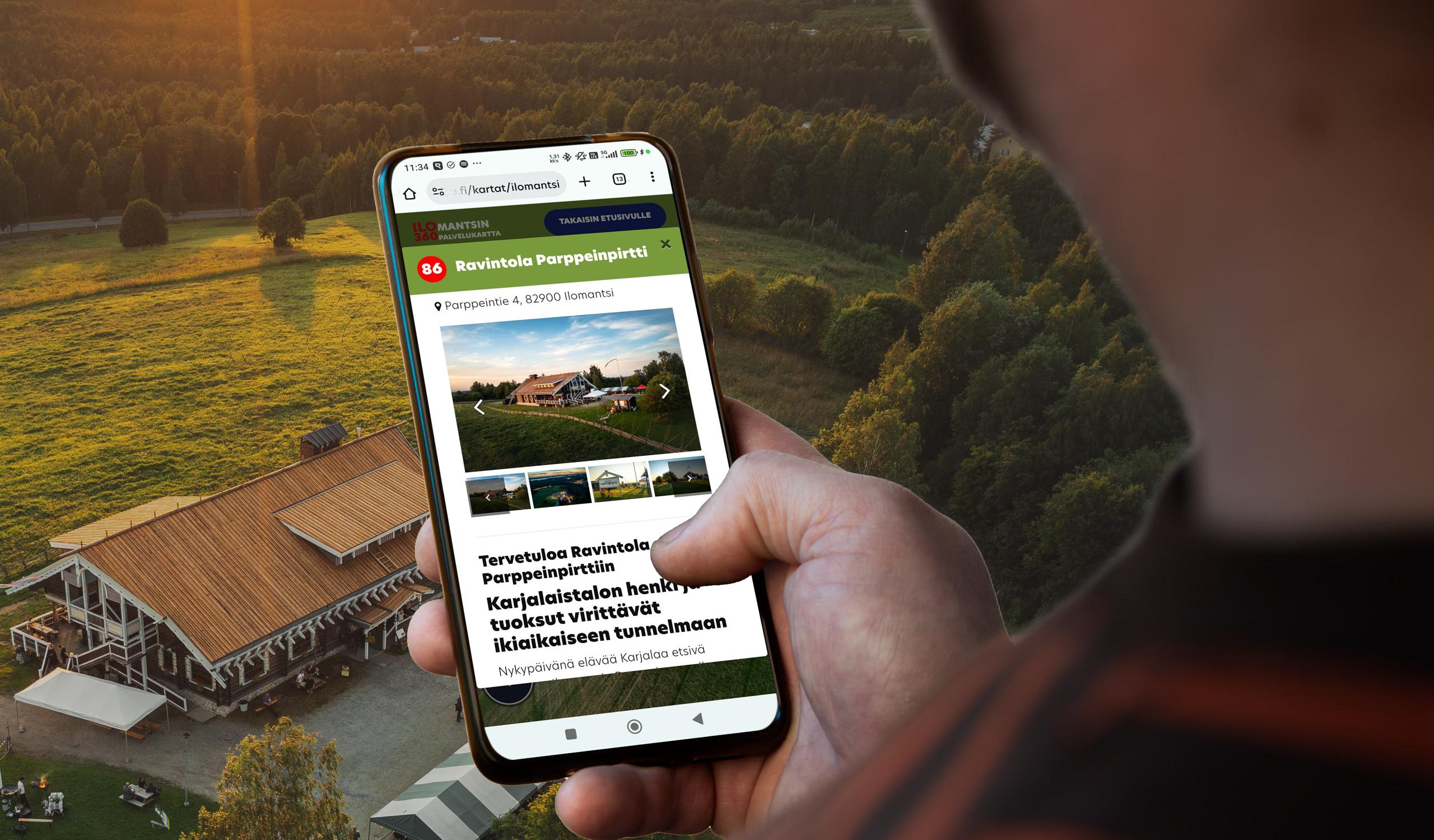 Henkil&ouml; k&auml;ytt&auml;&auml; &auml;lypuhelimella Ilomantsin 360 Palvelukarttaa, jonka n&auml;yt&ouml;ll&auml; on Ravintola Parppeinpirtin infokortti. Taustalla on ilmakuva samasta ravintolasta ja sit&auml; ymp&auml;r&ouml;iv&auml;st&auml; vaara- ja j&auml;rvimaisemasta auringonlaskun aikaan.