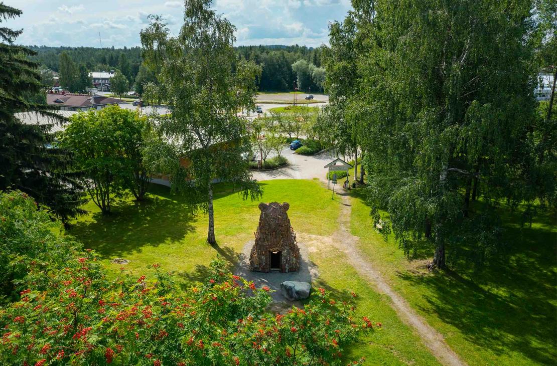 Ilmakuva suuresta, puusta tehdystä karhupatsaasta, ILOKARHU -nimisestä veistoksesta Ilomantsista. Karhu on koottu useista pienemmistä puupaloista ja laudoista. Kuvassa näkyy selvästi karhun karkea pintakerros. Taustalla on Ilomantsin tori.