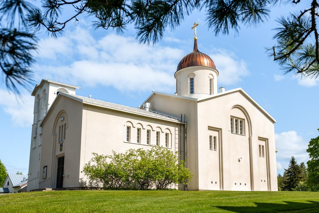 Valamon luostarin vaalea kirkkorakennus, jossa on kuparinen sipulikupoli ja ortodoksinen risti, kuvattuna alaviistosta aurinkoisena päivänä. Etualalla on vihreä nurmikko, ja kuvan yläosaa kehystävät männynoksat.