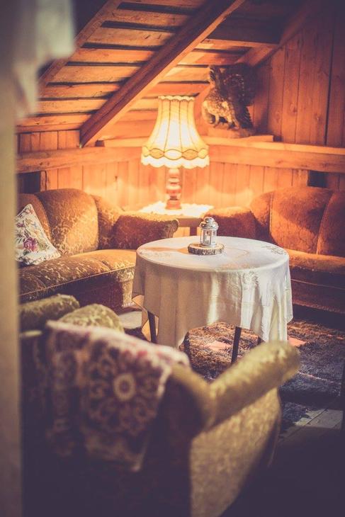 Kuva esittää Pappila Marjamäen ullakolla tai parvella sijaitsevaa oleskelutilaa. Tila on sisustettu kodikkaasti retrotyylisillä, sammaleenvihreillä tai oranssinruskeilla sohvilla ja nojatuoleilla. Huoneessa on puupaneeliseinät ja -katto. Keskellä on pyöreä pöytä valkoisella pöytäliinalla, ja tila on valaistu lämpimästi vanhanaikaisella pöytävalaisimella. Taustalla näkyy kattorakenteita ja ylätasolle vievät tikkaat.