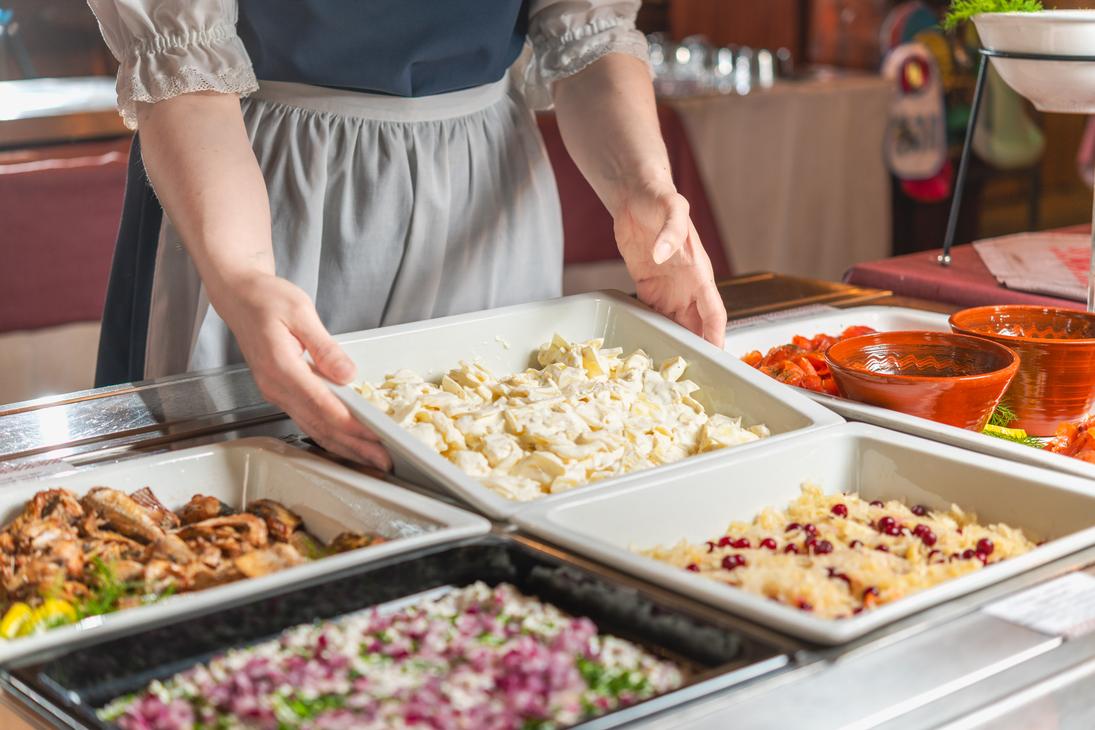 Lähikuva perinneasuun pukeutuneen tarjoilijan käsistä, jotka asettavat salaattivuokaa buffetpöytään Ravintola Parppeinpirtissä Ilomantsin Parppeinvaaralla. Pöydällä on tarjoiluastioissa erilaisia kylmiä ruokalajeja, kuten kalaa, pikkelöityä punasipulia ja hapankaalia puolukoilla.