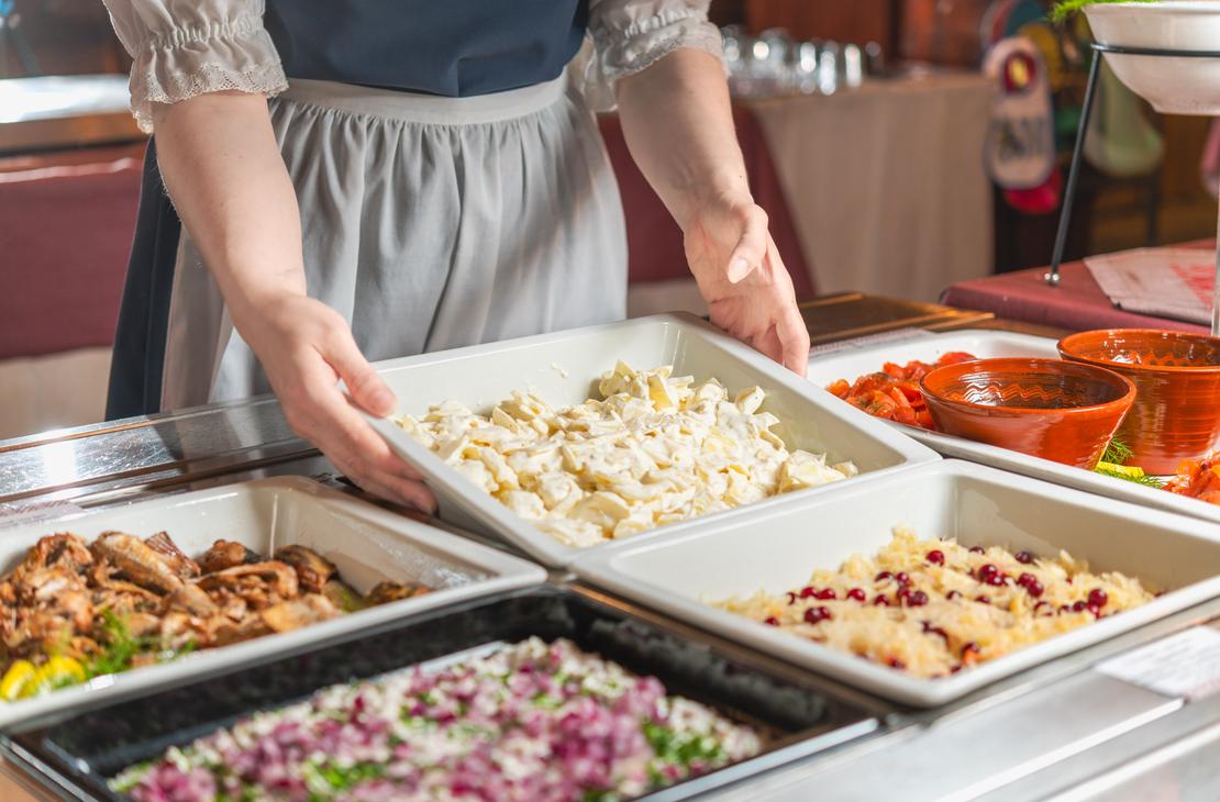 Lähikuva perinneasuun pukeutuneen tarjoilijan käsistä, jotka asettavat salaattivuokaa buffetpöytään Ravintola Parppeinpirtissä Ilomantsin Parppeinvaaralla. Pöydällä on tarjoiluastioissa erilaisia kylmiä ruokalajeja, kuten kalaa, pikkelöityä punasipulia ja hapankaalia puolukoilla.