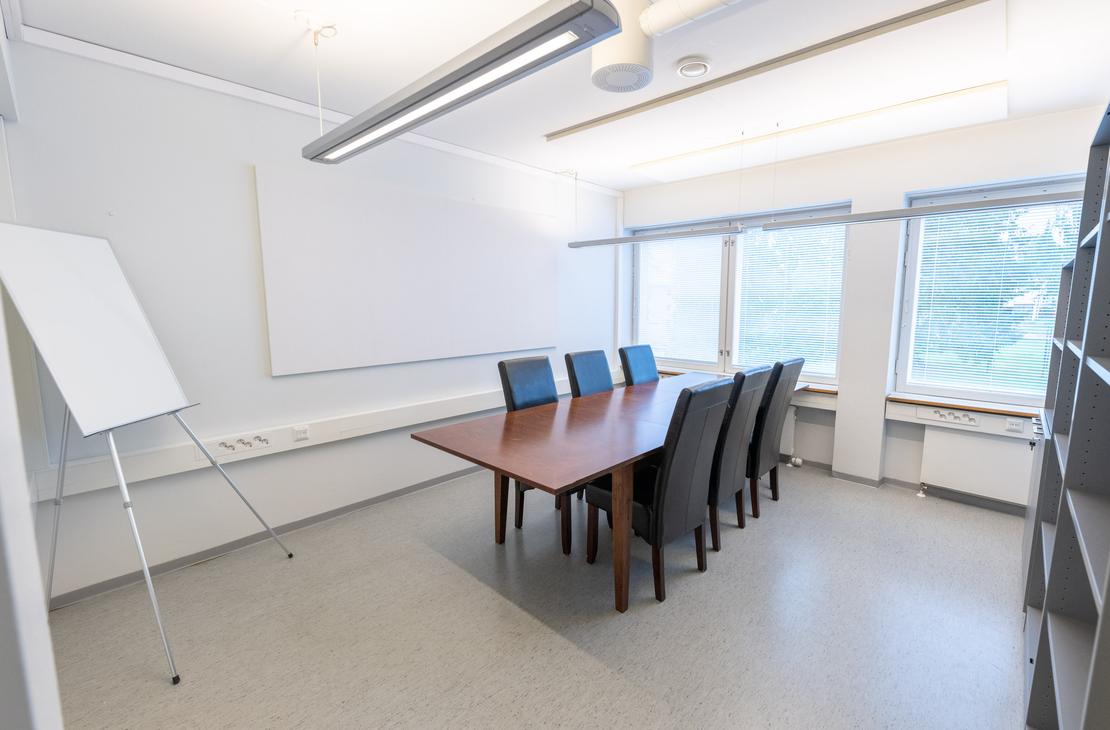 Kuva esitt&auml;&auml; Liikekulman Toimistohotellin pient&auml; neuvotteluhuonetta. Huoneessa on vaaleanharmaa lattia ja valkoiset sein&auml;t. Keskell&auml; on tummanruskea neuvottelup&ouml;yt&auml; ja sen ymp&auml;rill&auml; mustat nahka- tai kangasverhoillut tuolit. Vasemmalla on valkoinen fl&auml;ppitaulu ja suuri valkoinen sein&auml;taulu/valkokangas. Ikkunoista n&auml;kyy ulos vihre&auml;&auml;. Oikealla n&auml;kyy harmaa hyllykk&ouml;.