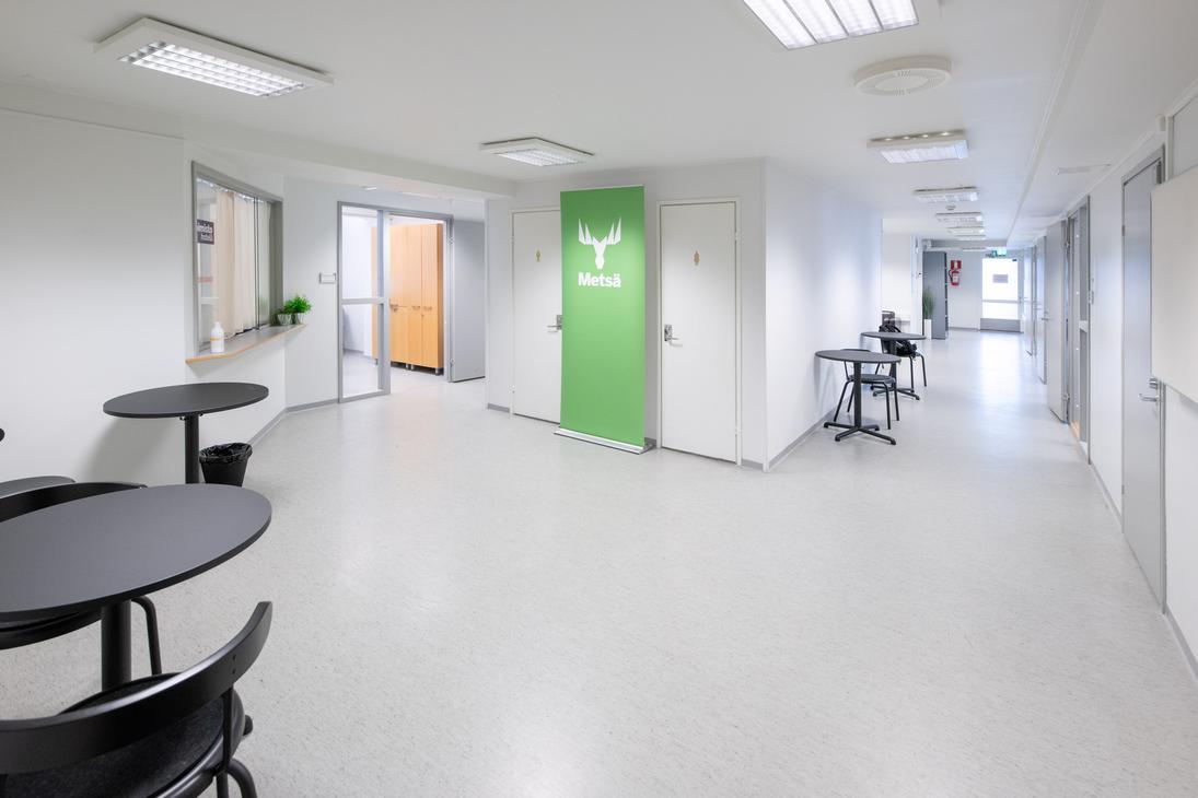 uva esittää Liikekulman Toimistohotellin modernia ja valoisaa käytävää tai aulatilaa. Käytävässä on vaalea lattia ja valkoiset seinät. Vasemmalla etualalla on pyöreitä, mustia pöytiä tuoleineen. Keskellä näkyy kirkkaan limevihreä ovi, jossa on logo (mahdollisesti yrityksen tunnus), ja taaempana oranssin ja keltaisen sävyisiä oviaukkoja. Oikealla on pitkä, valkoinen käytävä, jossa on työpöytiä.