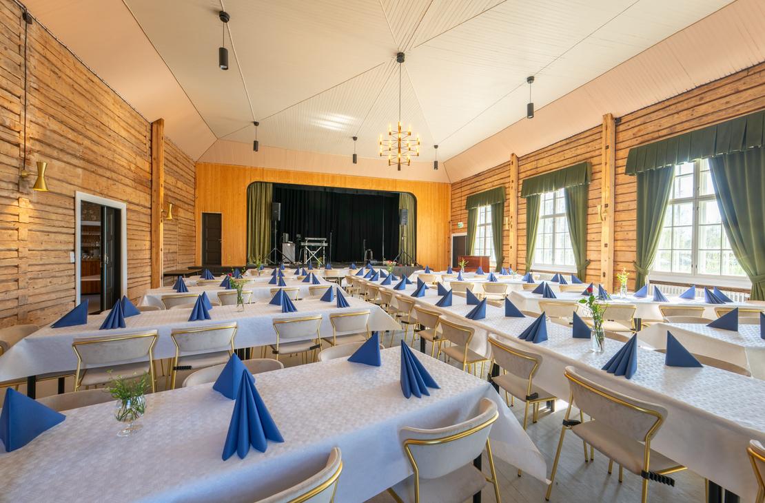 Kuva esittää Matkailukeskus Puu-Juuka Oy:n suurta juhla- tai ravintolasalia pitkien lounas- tai illallispöytien asetelmalla. Salissa on vaaleat puupaneeliseinät ja vaalea katto, josta riippuu riippuvalaisimia. Pöydät on katettu valkoisilla pöytäliinoilla ja niillä on taiteltuja sinisiä lautasliinoja. Salin päädyssä on esiintymislava keltaisella seinällä ja tummilla verhoilla. Ikkunoissa on tummanvihreät verhot.