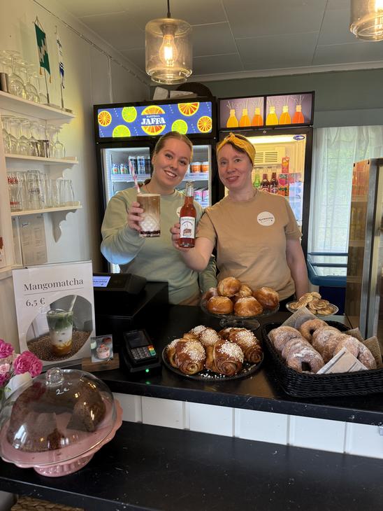 Kaksi hymyilevää työntekijää Satamakahvila-ravintola Kermacotan tiskillä. Toisella työntekijällä on käsissään pullollinen juomaa ja toisella täytetty lasi. Tiskin päällä on esillä houkuttelevia, vastaleivottuja korvapuusteja ja muita leivonnaisia koreissa. Taustalla näkyy kylmäkaappeja juomille.