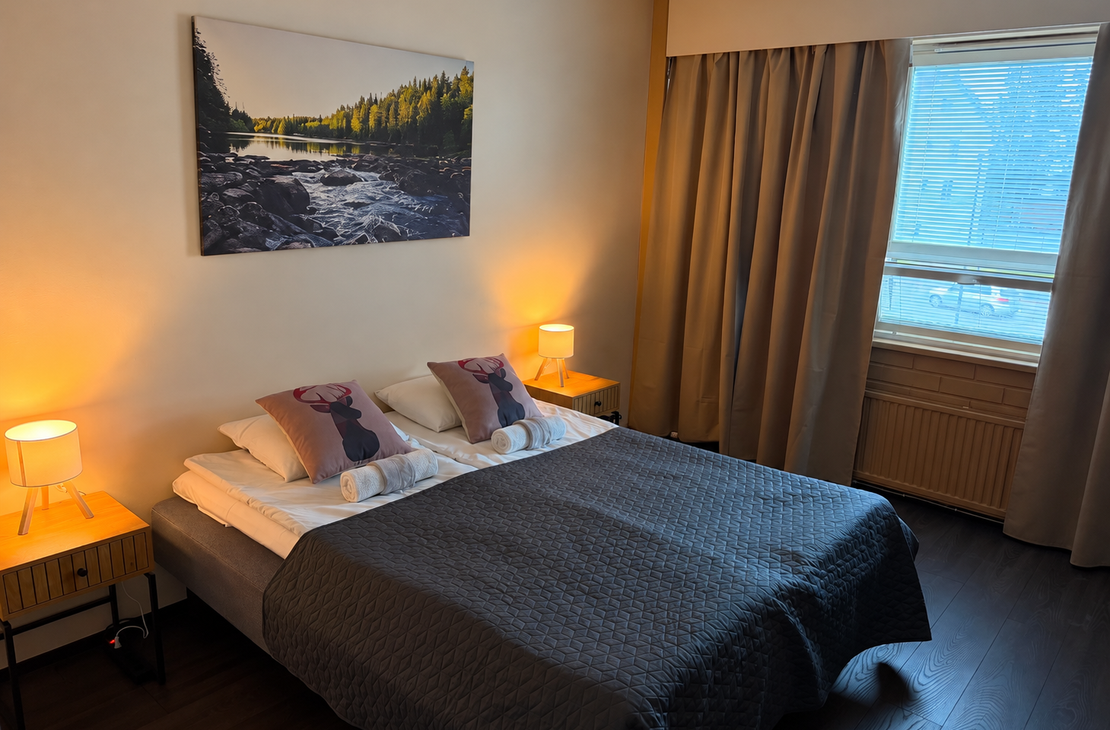 Viihtyisä hotellihuone, jossa on parivuode tummanharmaalla päiväpeitteellä, peuratyyneillä ja rullatuilla pyyhkeillä. Yöpöydillä hehkuvat lämpimät lamput, ja sängyn yläpuolella on suuri luontoaiheinen taulu virtavasta joesta.