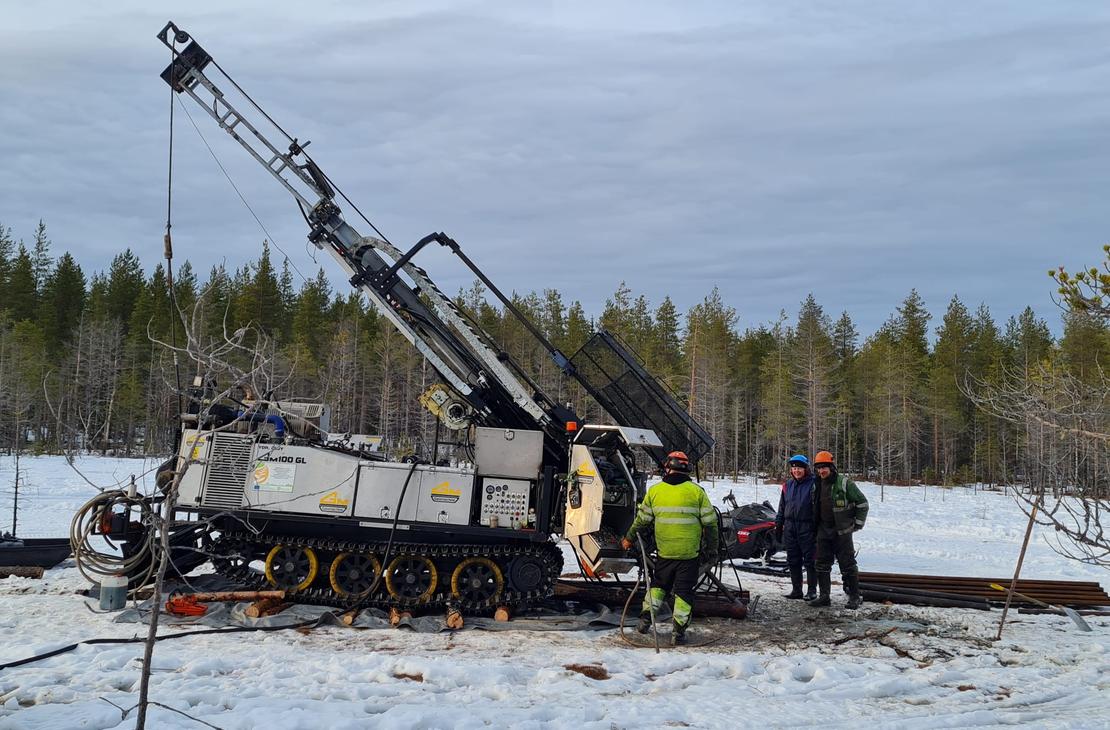 Kairaaja ja kaksi muuta henkilöä seisovat suuren telaketjuilla kulkevan kairauskoneen vieressä lumisella suolla. Taustalla on havumetsää ja pilvinen taivas.