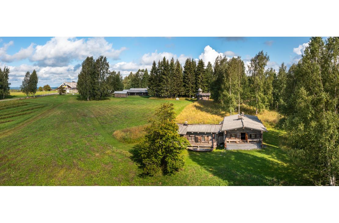  Laaja panoraamakuva, joka esitt&auml;&auml; Rajakenraalin majaa, perinteist&auml; hirsim&ouml;kki&auml; tai -rakennusta, joka sijaitsee avoimessa, kumpuilevassa maastossa. Rakennus on keskell&auml; oikealla ja sen edess&auml; on vihre&auml; nurmikentt&auml;. Alueella on runsaasti puita, erityisesti koivuja ja havupuita, ja taustalla avautuu maisema, jossa on lis&auml;&auml; rakennuksia ja peltoa. Taivas on sininen ja pilvinen.