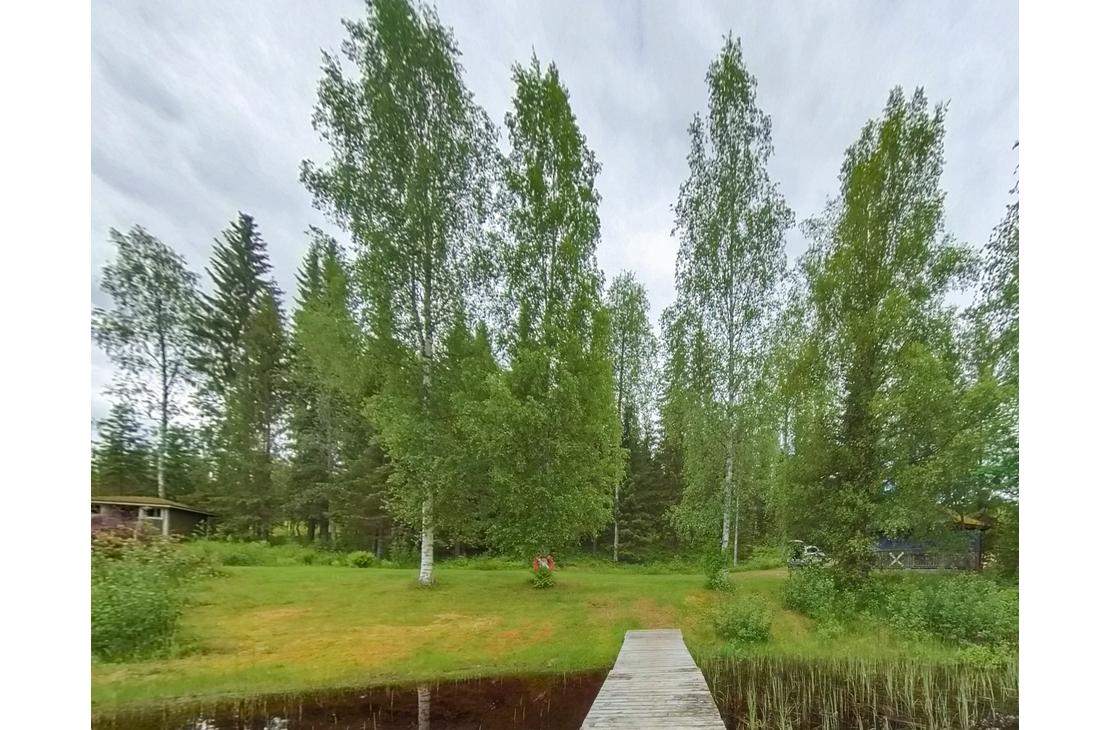 Kuva esitt&auml;&auml; kes&auml;ist&auml; maisemaa, jossa puinen laituri ulottuu etualalla tummaan veteen. Laiturilta avautuu n&auml;kym&auml; vihre&auml;lle nurmialueelle, jota reunustavat korkeat, hoikat koivut ja taempana tihe&auml;mpi sekamets&auml;. Taivas on pilvinen, ja nurmikolla koivujen v&auml;liss&auml; n&auml;kyy pieni, ruskea rakennelma tai esine.