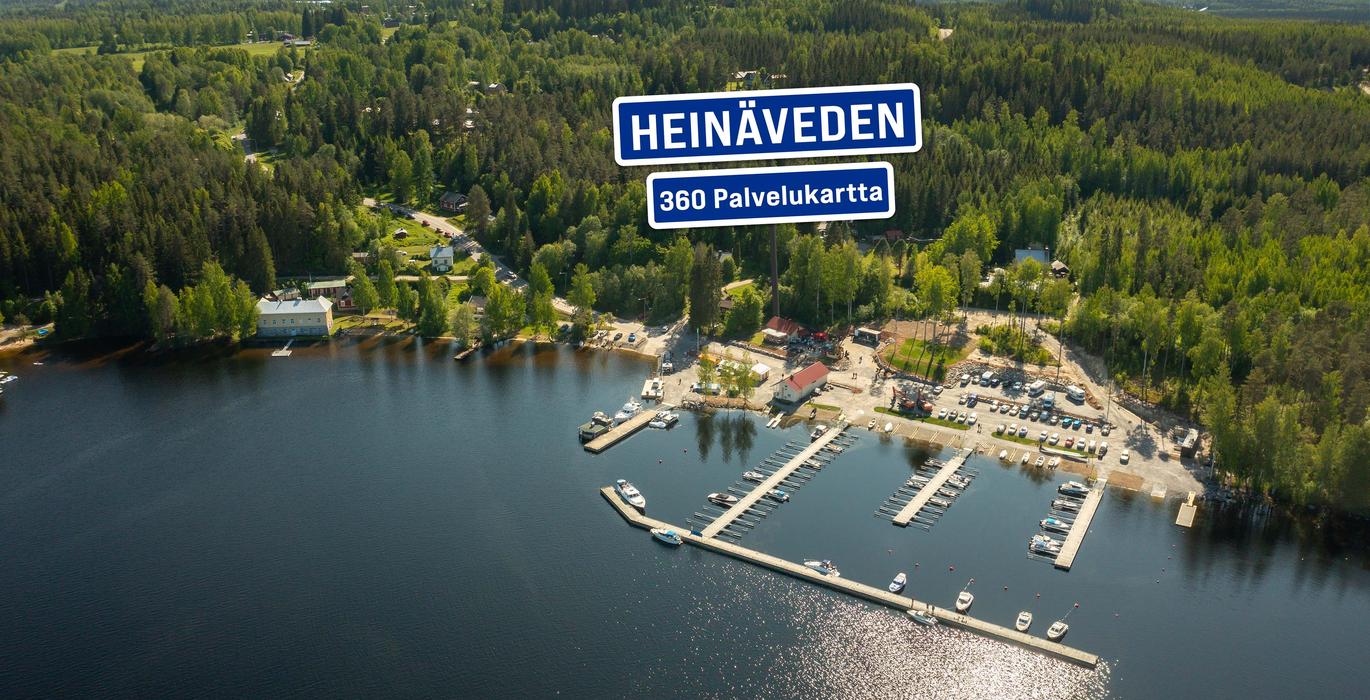 Ilmakuva Heinäveden satamasta, jossa on venelaitureita ja pysäköintialue järven rannalla. Taustalla näkyy laaja, metsäinen vaaramaisema. Kuvan päällä on sininen kyltti "HEINÄVEDEN 360 Palvelukartta".